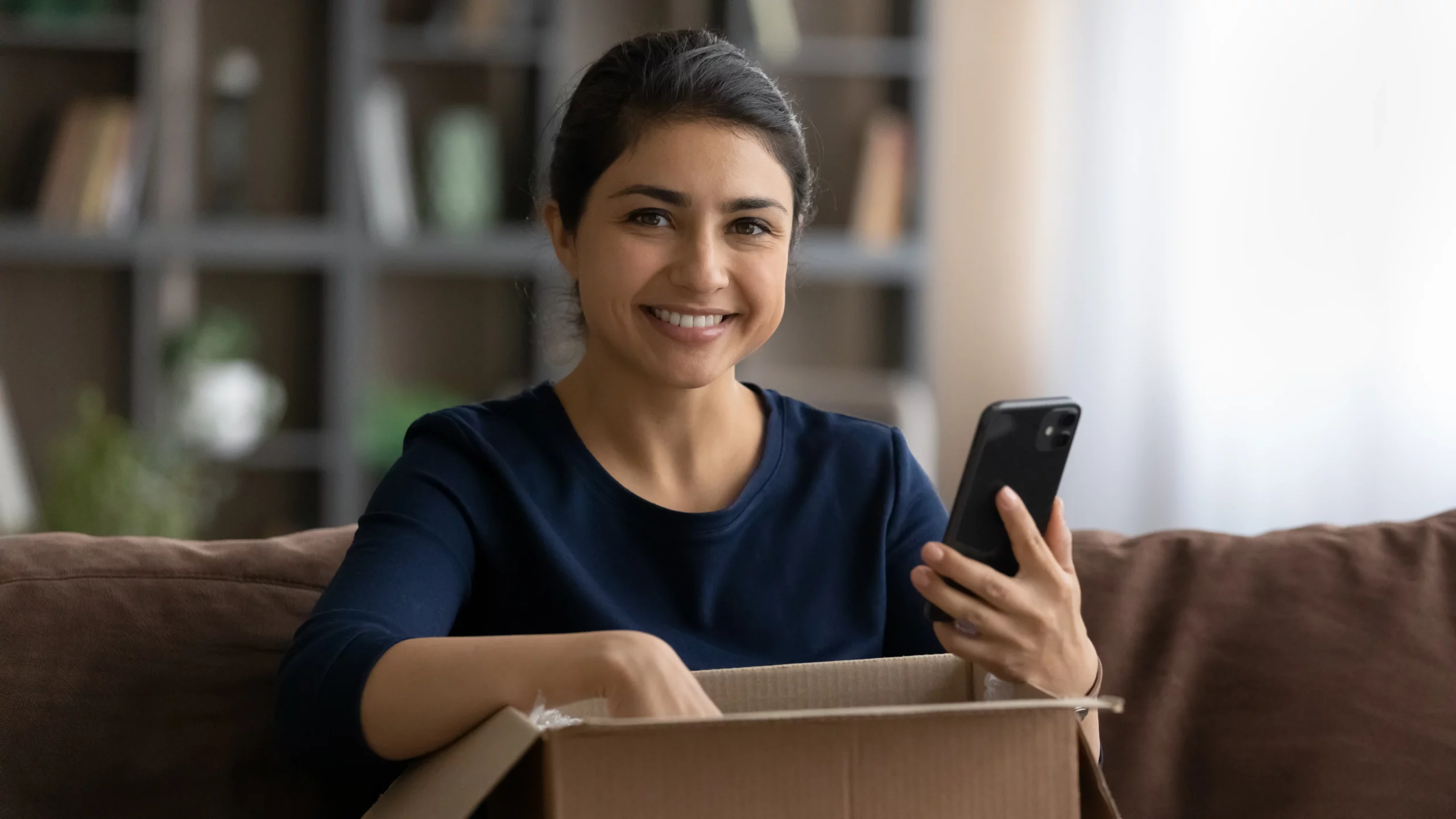 mujer sonriente sosteniendo un teléfono con la otra mano dentro de una caja de cartón abierta